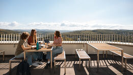 Drei Studentinnen sitzen am Tisch auf einer Terrasse mit Panoramablick.