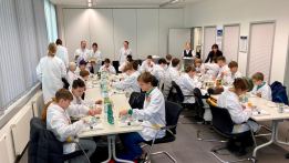 Schülerinnen und Schüler in weißen Kitteln sitzen und stehen an Tischen in einem großen Raum