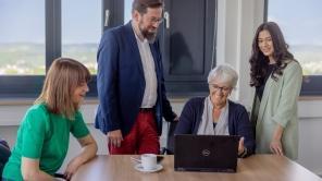 Vier Personen nehmen an einem Meeting teil. Ein Mann und eine Frau stehen links und rechts hinter einer weiteren Frau, die ihnen etwas am Laptop zeigt