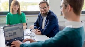 Drei Personen sitzen bei einer Besprechung, die Person im Vordergrund schaut auf einen Laptop