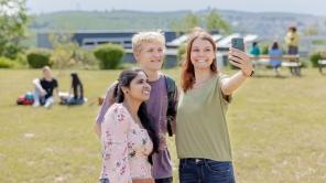 Studierende sitzen auf der LEOwiese und machen zu dritt ein Selfie.