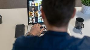 Person von hinten bei einer Videokonferenz mit Laptop. Daneben eine Kaffeetasse und Pflanze auf einem Schreibtisch