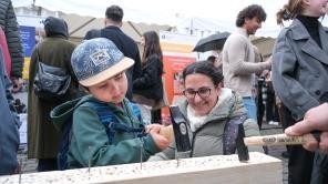 An einem Stand der Offenen Uni 2025 schlägt ein Junge mit einem Hammer einen Nagel in ein Brett. Um ihn herum laufen viele Menschen.