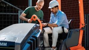 Studierender mit Bauhelm sitzt auf einem Arbeitsgerät und bekommt dieses erklärt