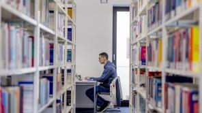 Ein Student sitzt lernend an einem Schreibtisch, umgeben von Bücherregalen in der Uni-Bibliothek.