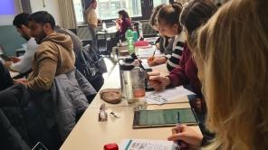 Studierende sitzen in einem Hörsaal an Tischen, schreiben und nutzen Laptops und Tablets während eines Vortrags