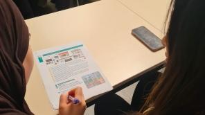 Zwei Studierende sitzen an einem Tisch, eine schreibt auf einem Blatt mit Text und Grafiken.