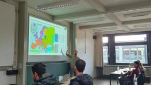 Dozent steht vor einer Leinwand mit einer farbigen Europakarte und zeigt darauf.