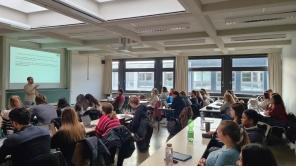 Vorlesungssaal mit Studierenden, die einem Vortrag folgen, während ein Dozent vor einer Projektion spricht.