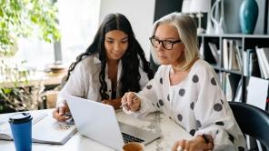 Zwei weiblich gelesene Personen sitzen zusammen am Schreibtisch und gucken gemeinsam in einen Laptop.