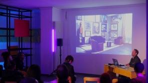 Vortragender sitzt an einem Tisch mit Laptop vor einer Wandprojektion eines Raums mit Sesseln und Bildern, Publikum sitzt im dunklen Raum