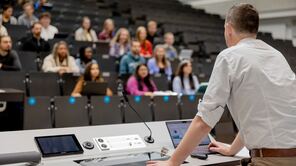 Dozent steht am Pult in einem Hörsaal und spricht zu Studierenden, die in aufsteigenden Sitzreihen zuhören.