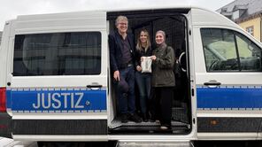 Prof. Kastenmüller posiert mit zwei seiner Mitarbeiterinnen in der Tür des Gefangenentransporters; die studentische Hilfskraft zeigt einen "Knastbeutel"