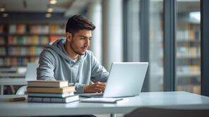 Junger Mann, der vor einem Laptop sitzt und neben dem ein Bücherstapel liegt