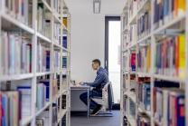 Ein Student sitzt lernend an einem Schreibtisch, umgeben von Bücherregalen in der Uni-Bibliothek.
