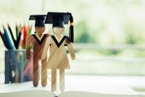 Zwei Holzfiguren mit Doktorandenhüten stehen vor einem Behälter mit Stiften.