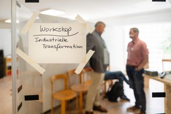 An der Glastür eines Seminarraums hängt ein Schild mit der Aufschrift &quot;Workshop Industrielle Transformation&quot;. Dahinter stehen unscharf erkennbare Personen.