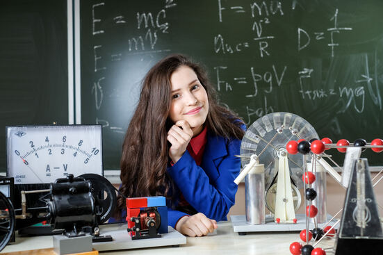 Eine Schülerin sitzt an einem Tisch mit Experimentiermaterialien, im Hintergrund ist eine Tafel mit Formeln zu sehen