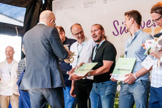 : Sven Bittenbinder (mittig, schwarzes Oberteil, Jeans) vom Lehrstuhl Wirtschaftsinformatik, insb. IT für die alternde Gesellschaft, der Universität Siegen, nimmt stellvertretend für das Projektkonsortium von KIWi@work den dritten Stern des REGIONALE 25 Förderprogramms entgegen.