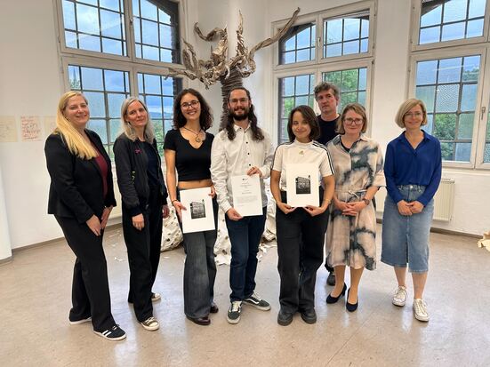 Drei Kunstreisträger*innen halten ihre Urkunde hoch. Neben ihnen stehen Kanzlerin Iris Litty sowie Professor*innen aus dem Institut Kunst.