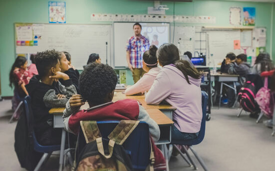 Schulklasse: Schüler sitzen an Tischen, vorn steht ein Lehrer