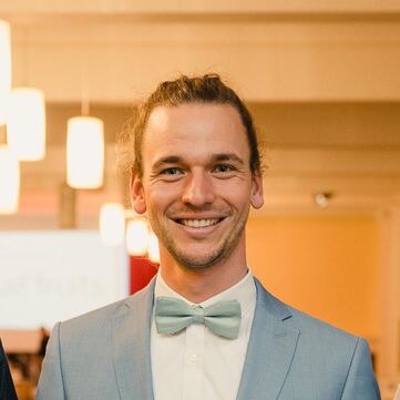 Philipp Spelten, hellblauer Anzug, hellgrüne Fliege, lange Haare im Zopf, breites Lächeln
