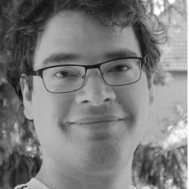 Ein Portraitfoto einer Person mit kurzen, schwarzen, leicht lockigen Haaren und einer Brille