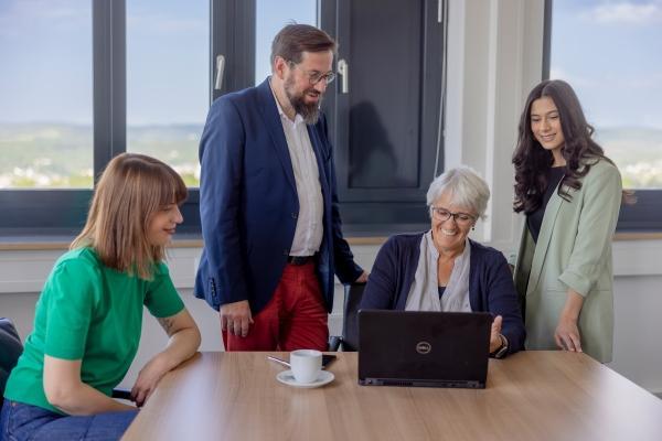 Vier Personen nehmen an einem Meeting teil. Ein Mann und eine Frau stehen links und rechts hinter einer weiteren Frau, die ihnen etwas am Laptop zeigt