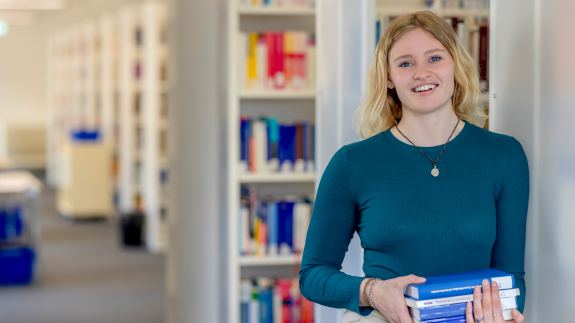 Studentin steht mit Buch in der Hand vor Bücherregal