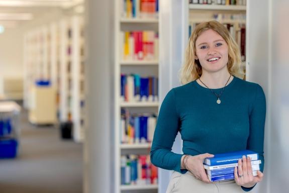 Studentin steht mit Buch in der Hand vor Bücherregal