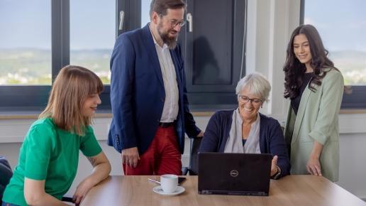 Vier Personen nehmen an einem Meeting teil. Ein Mann und eine Frau stehen links und rechts hinter einer weiteren Frau, die ihnen etwas am Laptop zeigt