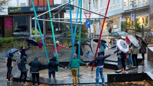 Eine Gruppe spielt: Menschen stehen im Kreis um eine moderne Statue herum und halten sich an der Hand.