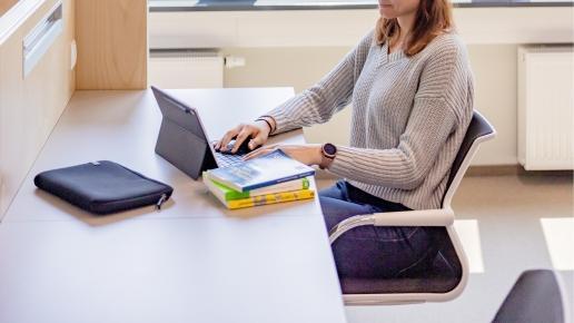 Studentin sitzt an Schreibtisch vor geöffnetem Laptop und Bücherstapel