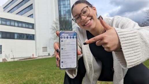 Person zeigt mit Finger auf Smartphone, darauf ist die Uni Siegen App zu sehen