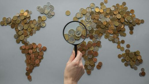 Weltkarte aus Münzen, eine Hand mit Lupe richtet sich auf Europa.