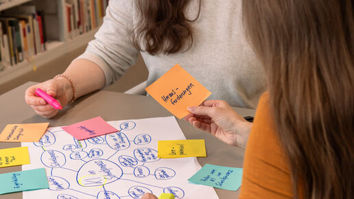 Zwei Studierende sitzen an einem Tisch zusammen. Vor ihnen liegen Arbeitsmaterialien: ein Blatt mit einer Mind Map, Klebezettel und Textmarker