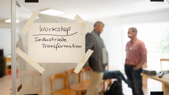An der Glastür eines Seminarraums hängt ein Schild mit der Aufschrift &quot;Workshop Industrielle Transformation&quot;. Dahinter stehen unscharf erkennbare Personen.
