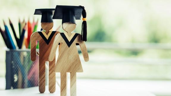 Zwei Holzfiguren mit Doktorandenhüten stehen vor einem Behälter mit Stiften.