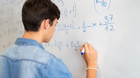Junge von Hinten schreibt mit einem Stift an einer Tafel auf der mathematische Terme zu sehen sind.