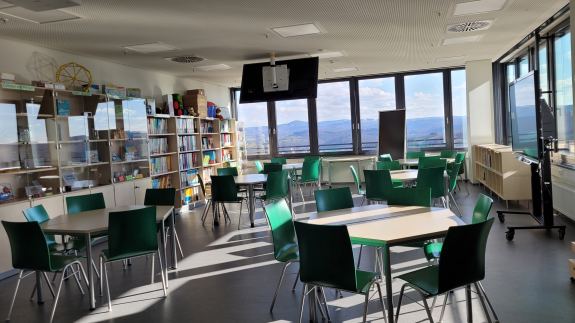 Ein heller, großer Raum mit Gruppentischen, grünen Stühlen, einer großen Fensterfront mit Blick auf eine Hügellandschaft und Regalen voller Lernmaterialien.