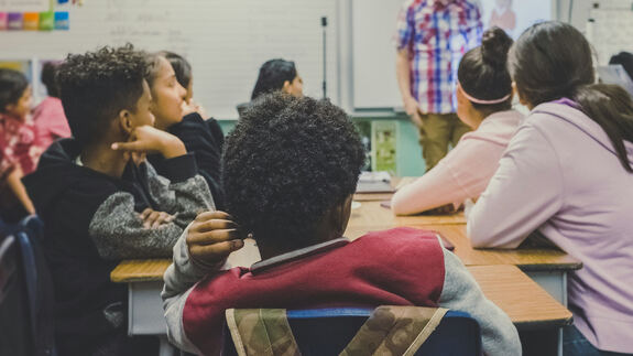 Schulklasse: Schüler sitzen an Tischen, vorn steht ein Lehrer
